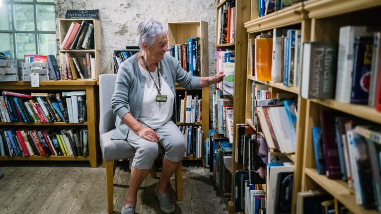 Lady with short grey hair and a black volunteer lanyard and name badge sitting on a chair in Sizergh's second hand bookshop. She is smiling and picking out a book.